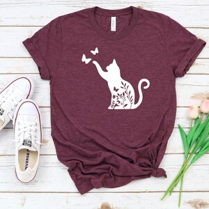 a burgundy t-shirt with a white cat silhouette and butterflies printed on it, along with a pair of white sneakers and a bouquet of pink flowers.