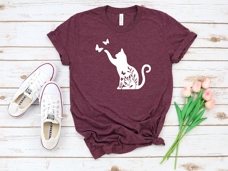a burgundy t-shirt with a white cat silhouette and butterflies printed on it, along with a pair of white sneakers and a bouquet of pink flowers.