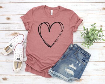 May include: A dusty rose t-shirt with a black heart design and the text "Your text". The shirt is paired with denim shorts and white sneakers with red accents, all arranged on a light wood surface.