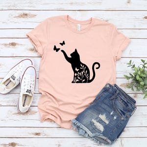 A pink t-shirt with a black cat silhouette and butterflies printed on it, along with a pair of denim shorts and white sneakers, all laid out on a wooden surface.