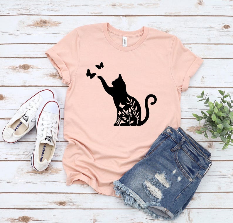 A pink t-shirt with a black cat silhouette and butterflies printed on it, along with a pair of denim shorts and white sneakers, all laid out on a wooden surface.
