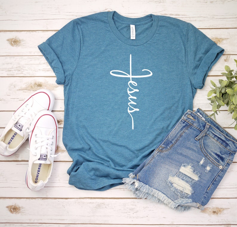 K&ouml;nnte beinhalten: Ein t&uuml;rkisfarbenes T-Shirt mit einem wei&szlig;en Kreuz und dem Wort "Jesus", das vertikal darauf geschrieben ist. Das Shirt liegt auf einer Holzoberfl&auml;che mit einem Paar wei&szlig;en Turnschuhen und einer Jeans-Shorts.
