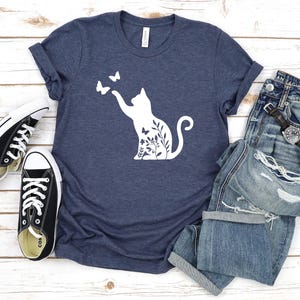 A casual outfit consisting of a blue t-shirt with a white cat silhouette, distressed jeans, and black and white sneakers is laid out on a wooden surface.