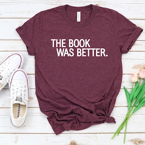 May include: A maroon t-shirt with the text "THE BOOK WAS BETTER" in white. The shirt is next to a pair of white trainers and a bouquet of pink tulips. The items are arranged on a light wood-grain surface.