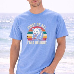 A man wearing a blue t-shirt with a graphic design and text printed on it, standing on a beach with the ocean in the background.