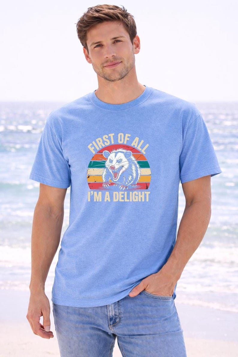A man wearing a blue t-shirt with a graphic design and text printed on it, standing on a beach with the ocean in the background.