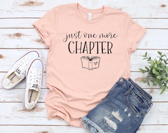 May include: A light pink t-shirt with the text "just one more CHAPTER" and a book graphic. The shirt is paired with denim shorts and white sneakers. The items are arranged on a light wood-grain surface.
