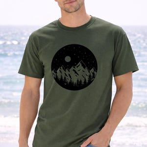 A man wearing a green t-shirt with a mountain landscape design stands on a beach, with the ocean visible in the background.