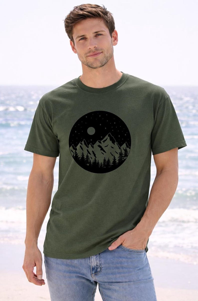 A man wearing a green t-shirt with a mountain landscape design stands on a beach, with the ocean visible in the background.