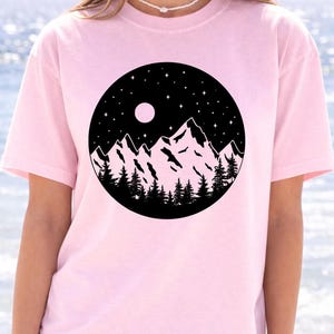 A woman wearing a pink t-shirt with a graphic design of a mountain landscape under a night sky.