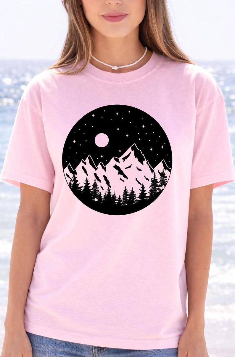 A woman wearing a pink t-shirt with a graphic design of a mountain landscape under a night sky.