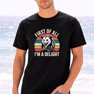 A man wearing a black t-shirt with a graphic of a smiling opossum and the text "First of all I'm a delight" is standing on a beach.