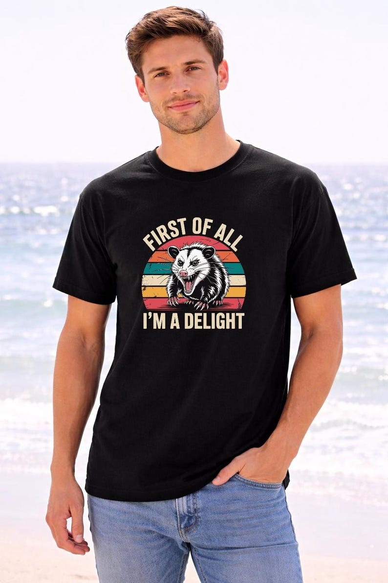 A man wearing a black t-shirt with a graphic of a smiling opossum and the text "First of all I'm a delight" is standing on a beach.