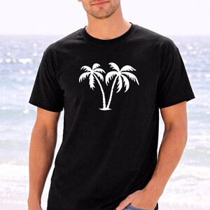 A man wearing a black t-shirt with a white palm tree graphic stands on a beach, with the ocean visible in the background.