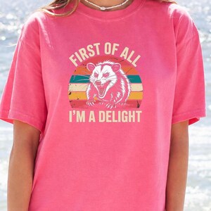A woman wearing a pink t-shirt with a graphic design of a smiling opossum and the text "First of all I'm a delight".
