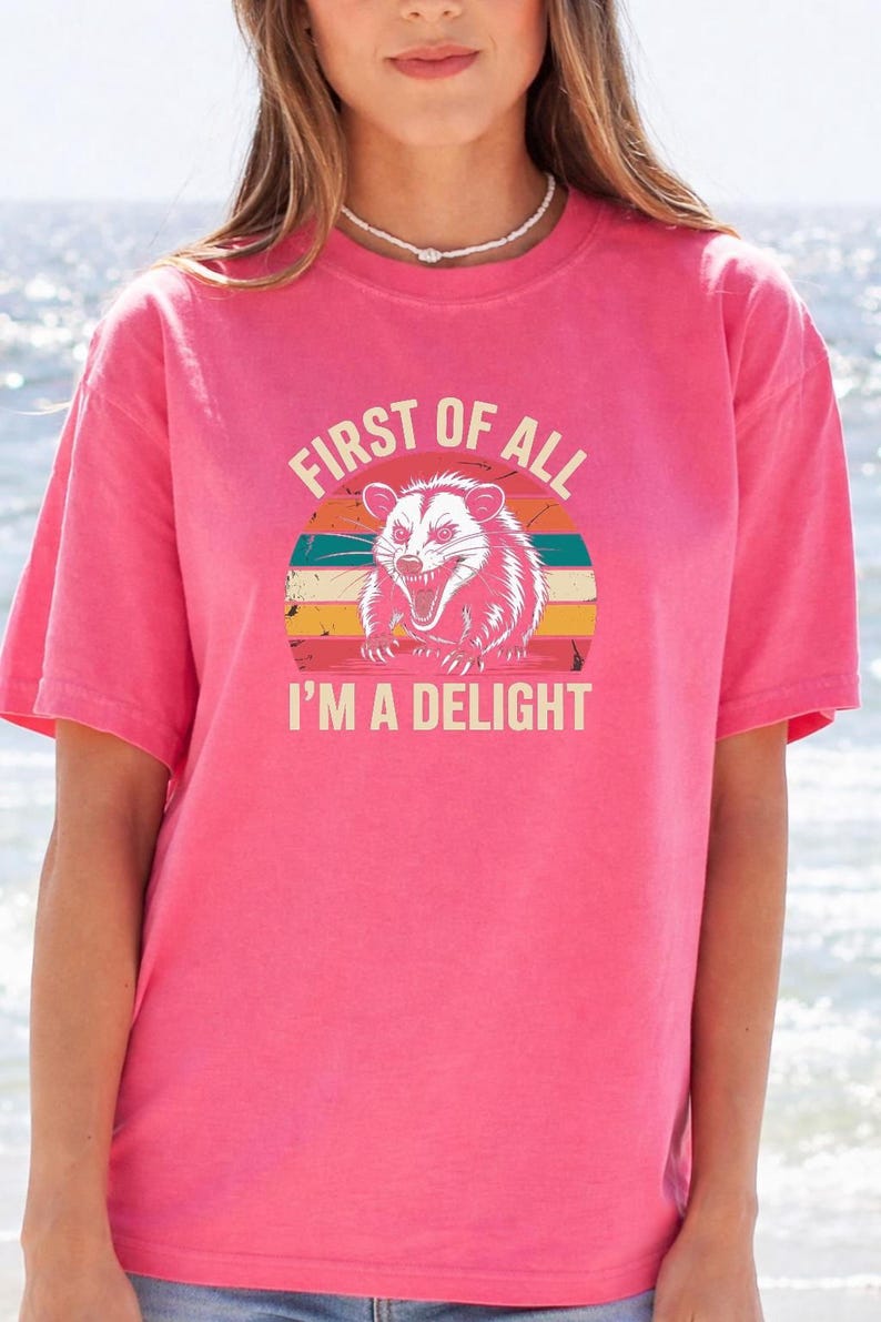 A woman wearing a pink t-shirt with a graphic design of a smiling opossum and the text "First of all I'm a delight".