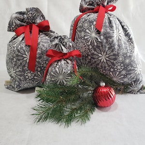 May include: Three gray fabric gift bags with white snowflake patterns and red ribbon ties. The bags are arranged on a white surface with a green pine branch and a red Christmas ornament.