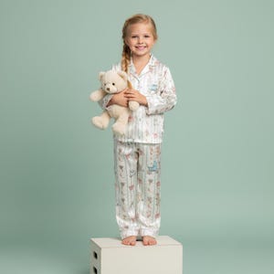 K&ouml;nnte beinhalten: Ein junges M&auml;dchen tr&auml;gt einen wei&szlig;en Pyjama mit vertikalem Streifenmuster und Illustrationen zum Thema Baby. Der Pyjama hat ein Oberteil mit Kragen und eine Hose. Sie h&auml;lt einen beigen Teddyb&auml;ren. Der Hintergrund ist einfarbig hellgr&uuml;n.