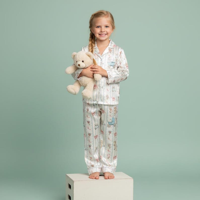 K&ouml;nnte beinhalten: Ein junges M&auml;dchen tr&auml;gt einen wei&szlig;en Pyjama mit vertikalem Streifenmuster und Illustrationen zum Thema Baby. Der Pyjama hat ein Oberteil mit Kragen und eine Hose. Sie h&auml;lt einen beigen Teddyb&auml;ren. Der Hintergrund ist einfarbig hellgr&uuml;n.