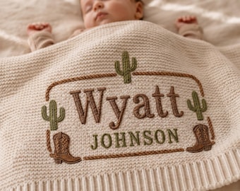 May include: A cream-colored knit baby blanket featuring an embroidered design with the name Wyatt Johnson in brown and green. The embroidery includes two green cacti, two brown cowboy boots, and a rope-style border framing the text.