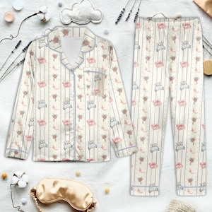 K&ouml;nnte beinhalten: Cremefarbenes Pyjama-Set mit einem sich wiederholenden Muster aus Autos, Blumenstr&auml;u&szlig;en und Geschenkboxen. Das lang&auml;rmlige Oberteil und die Hose haben vertikale blaue Streifen und blaue Paspeln. Eine goldfarbene Augenmaske ist ebenfalls sichtbar.