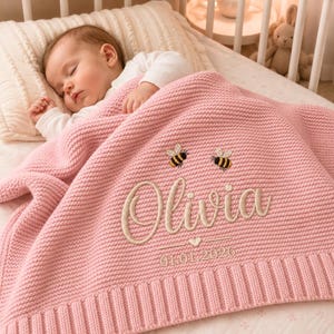 May include: A soft pink knitted baby blanket featuring an embroidered design of two small bees, the name Olivia in cursive script, a small heart icon, and the date 01.01.2026. The blanket has a textured garter stitch pattern and a ribbed border.