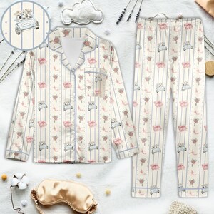 K&ouml;nnte beinhalten: Cremefarbenes Pyjama-Set mit einem sich wiederholenden Muster eines Oldtimers mit Blumen, rosa Schleifen und Blumenstr&auml;u&szlig;en. Das Set beinhaltet ein Hemd mit Kn&ouml;pfen und einer Brusttasche sowie eine passende Hose. Blaue Streifen und Verzierungen akzentuieren das Set.