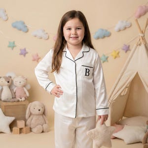 May include: A young person wearing white pajamas with black trim and a pocket with the letter "B". The pajamas are paired with a star-shaped pillow. The background includes a teepee, stuffed animals, and a garland of stars and clouds.