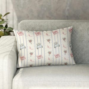 K&ouml;nnte beinhalten: Rechteckiges Zierkissen mit cremefarbenem Hintergrund und einem sich wiederholenden Muster aus hellblauen Streifen, rosa Geschenkboxen, Blumenstr&auml;u&szlig;en und Oldtimern. Das Kissen liegt auf einem grauen Sofa.