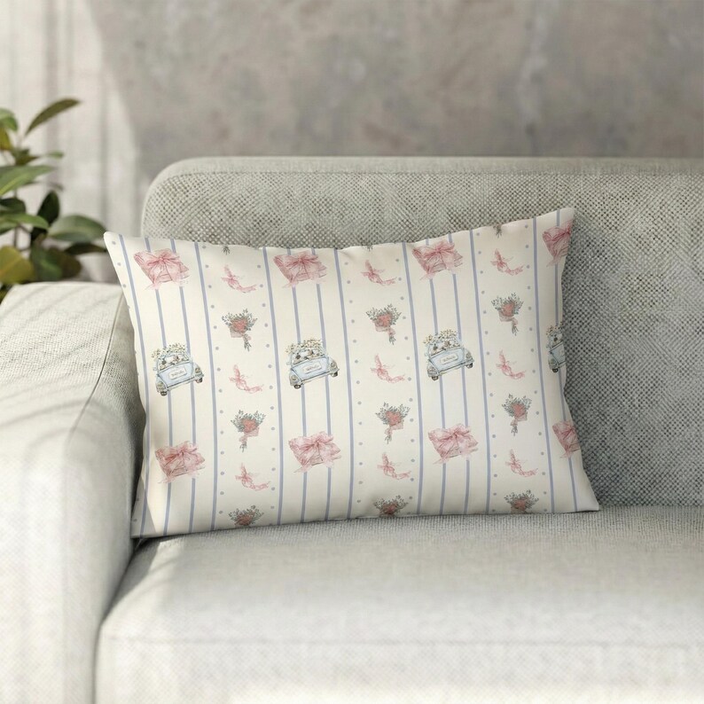 K&ouml;nnte beinhalten: Rechteckiges Zierkissen mit cremefarbenem Hintergrund und einem sich wiederholenden Muster aus hellblauen Streifen, rosa Geschenkboxen, Blumenstr&auml;u&szlig;en und Oldtimern. Das Kissen liegt auf einem grauen Sofa.