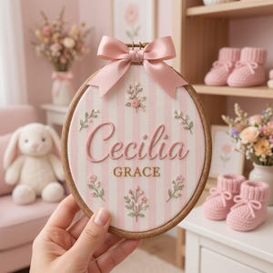 Może przedstawiać: Okrągły drewniany tamborek z tłem z tkaniny w jasnoróżowe i białe paski. Imię Cecilia jest wyhaftowane różową kursywą, a poniżej znajduje się imię GRACE złotymi literami. Małe gałązki kwiatowe otaczają tekst, a na górze przymocowana jest różowa satynowa kokardka.