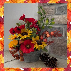 May include: A fall-themed floral arrangement in a galvanized metal bucket. The arrangement includes sunflowers, red poppies, berries, and greenery. A decorative ribbon and two pine cones complete the display. The background features a stone wall.