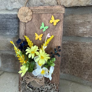 May include: A rustic wooden wall hanging with a jute rope handle and a burlap flower. The wooden plaque features three yellow and one green butterfly cutouts. A small pot of artificial flowers, including yellow daisies and white blossoms, is attached to the bottom.