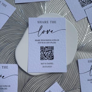 May include: White cards with black text that reads "Share the love" and includes a QR code. The text also says "Share your photos with us! Just scan and upload." and includes the date "May & Daniel 21.05.2025".