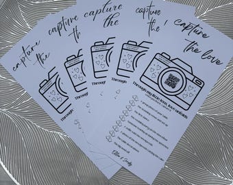 May include: Several light blue cards with the phrase "capture the love" and a camera illustration. They are placed on a silver charger plate with a leaf design. The text "Through my little lens, love unfolds" is also visible.