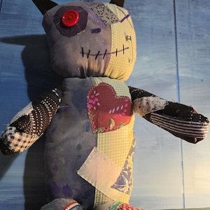 May include: A handmade plush doll constructed from various fabric patches in shades of blue, white, and black. The doll features a single red button eye, dark pointed ears, a stitched mouth, and a prominent heart-shaped patch on its chest.