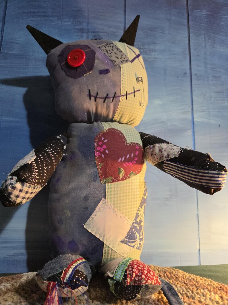 May include: A handmade plush doll constructed from various fabric patches in shades of blue, white, and black. The doll features a single red button eye, dark pointed ears, a stitched mouth, and a prominent heart-shaped patch on its chest.