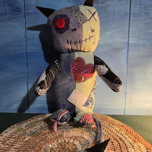 May include: A handmade fabric doll featuring a patchwork design with various patterns and textures. The doll has two pointed ears, a single red button eye, a stitched mouth, a safety pin detail, and a heart-shaped patch on its chest with a tail.
