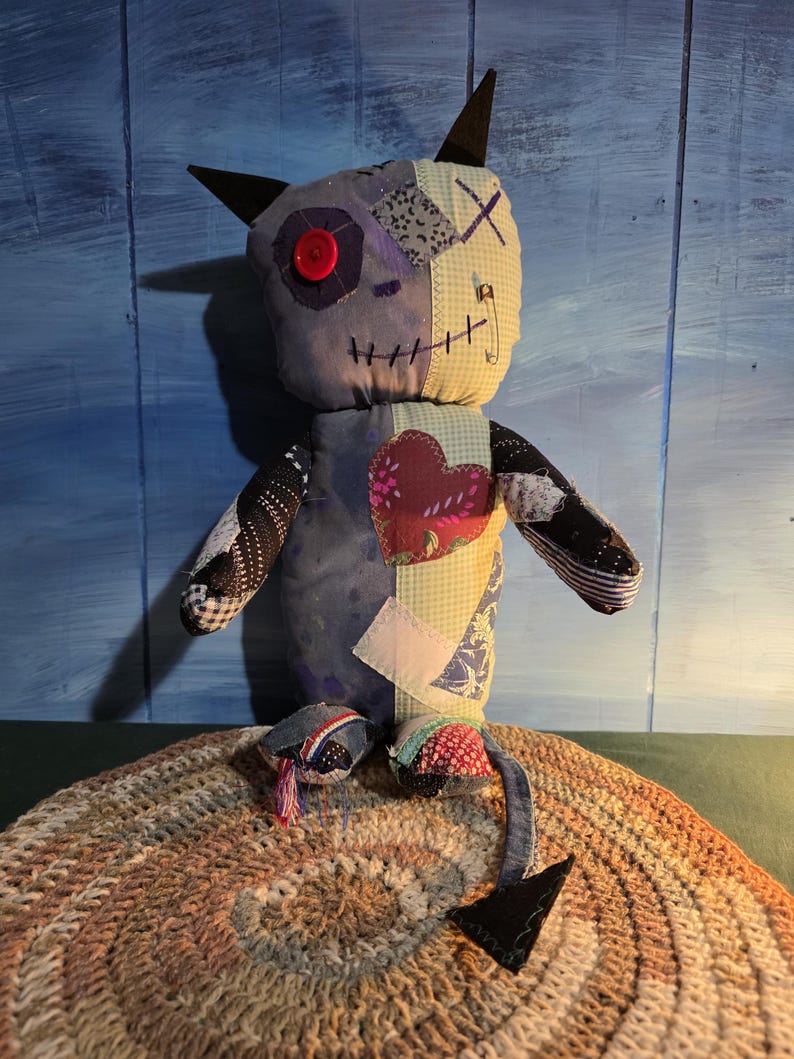 May include: A handmade fabric doll featuring a patchwork design with various patterns and textures. The doll has two pointed ears, a single red button eye, a stitched mouth, a safety pin detail, and a heart-shaped patch on its chest with a tail.