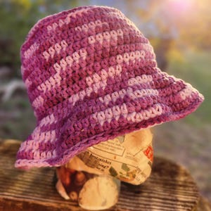 May include: A crocheted bucket hat in shades of pink and purple. The hat is made with a textured stitch and has a wide brim.