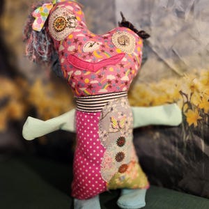 May include: A handmade fabric doll with a heart-shaped head and a patchwork dress. The head is pink with a whimsical design of stars, rainbows, and clouds. It has blue hair, a pink mouth, and decorative patches. The doll's dress is a mix of pink polka dots and floral patterns.