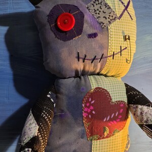 May include: A handmade plush doll featuring a patchwork design with a purple fabric face, a red button eye, and black felt ears. The body includes green gingham, black star-patterned fabric, and a large maroon heart patch with visible stitching.