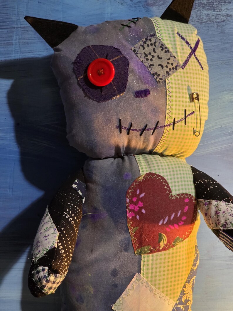 May include: A handmade plush doll featuring a patchwork design with a purple fabric face, a red button eye, and black felt ears. The body includes green gingham, black star-patterned fabric, and a large maroon heart patch with visible stitching.