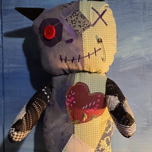 May include: A handmade plush doll featuring a patchwork design with various fabric patterns including light blue gingham, dark purple, and black star prints. It has a red button eye, a safety pin, and a large heart-shaped fabric patch on the chest.