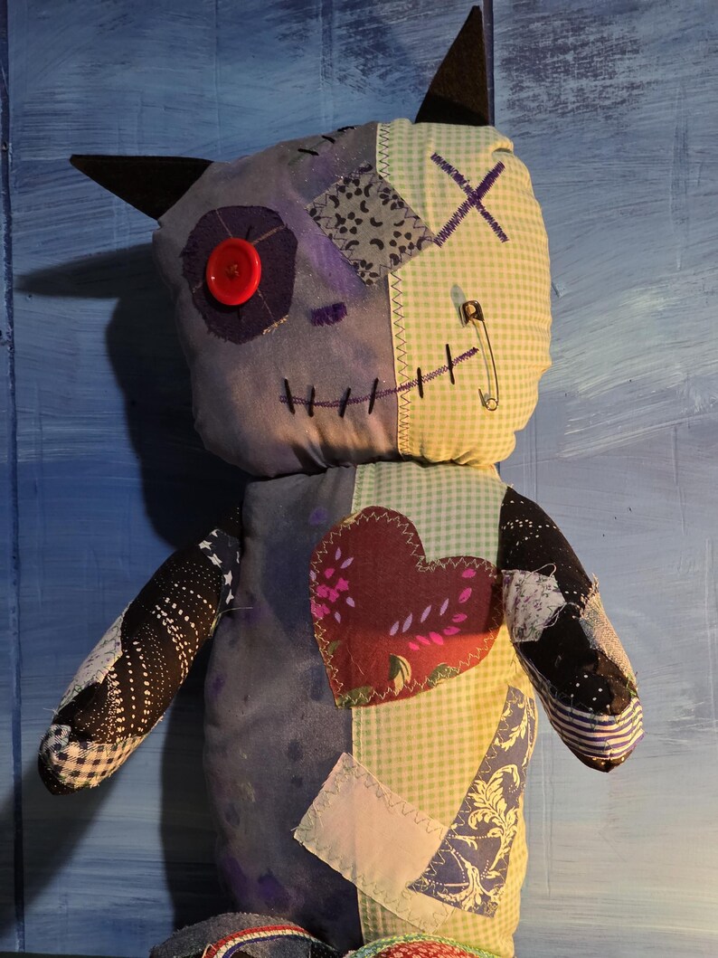 May include: A handmade plush doll featuring a patchwork design with various fabric patterns including light blue gingham, dark purple, and black star prints. It has a red button eye, a safety pin, and a large heart-shaped fabric patch on the chest.