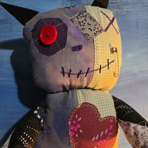 May include: A handmade plush doll featuring a patchwork design with blue, yellow, and black fabric. It has two pointed black ears, a single red button eye, a stitched mouth, and a large heart-shaped patch on the chest. A silver safety pin is attached to the side.