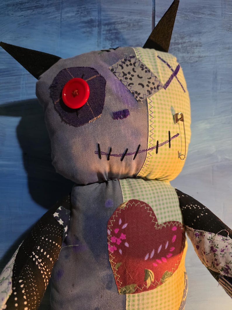 May include: A handmade plush doll featuring a patchwork design with blue, yellow, and black fabric. It has two pointed black ears, a single red button eye, a stitched mouth, and a large heart-shaped patch on the chest. A silver safety pin is attached to the side.