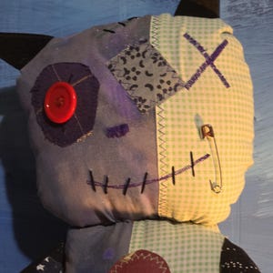 May include: A plush doll featuring a patchwork design with a blue fabric side and a light green gingham side. It has a red button eye, a safety pin, black stitched mouth details, and dark pointed ears. The chest area includes a dark heart patch.