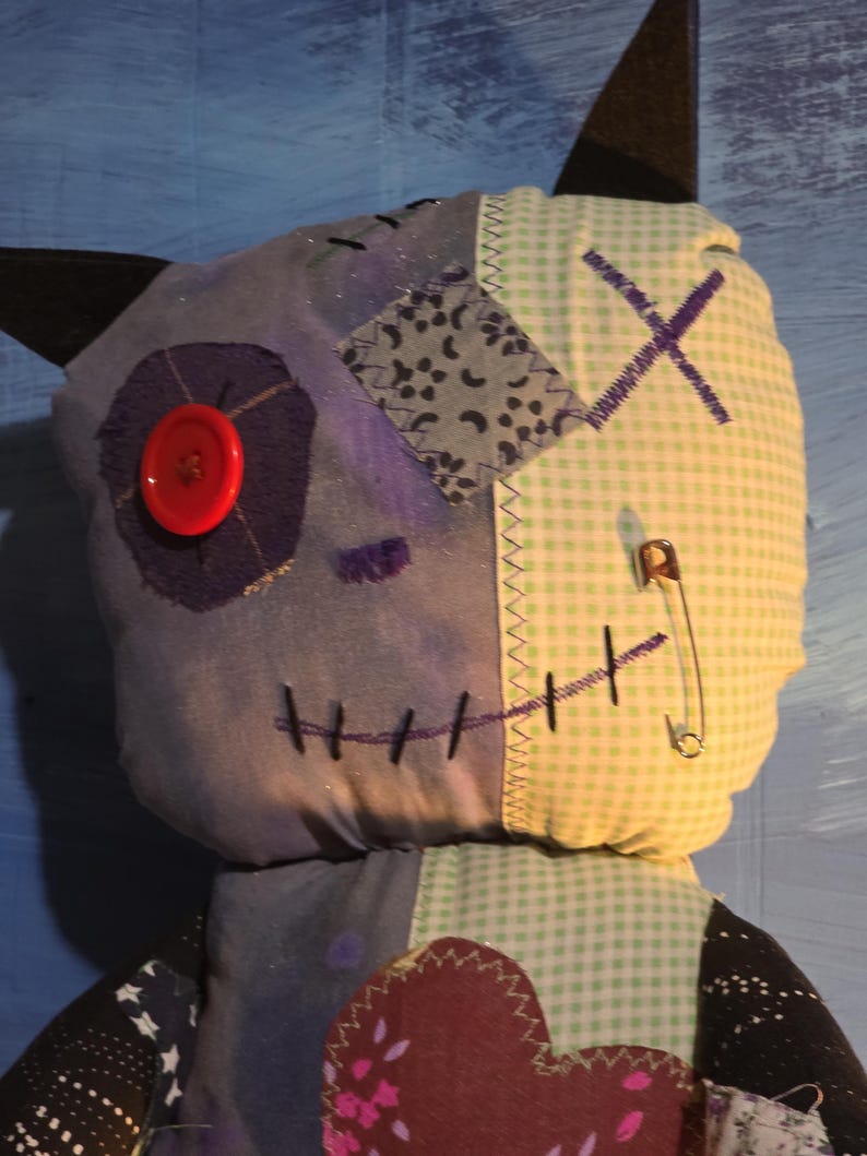 May include: A plush doll featuring a patchwork design with a blue fabric side and a light green gingham side. It has a red button eye, a safety pin, black stitched mouth details, and dark pointed ears. The chest area includes a dark heart patch.
