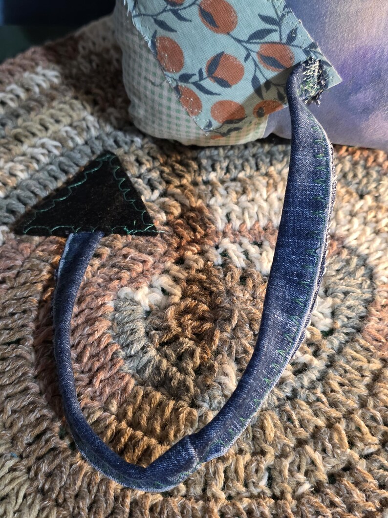 May include: A handcrafted plush toy tail made from blue denim fabric with visible green stitching along the edges. The tail features a triangular black felt tip at the end and is attached to a soft body covered in light blue fabric with an orange fruit pattern.